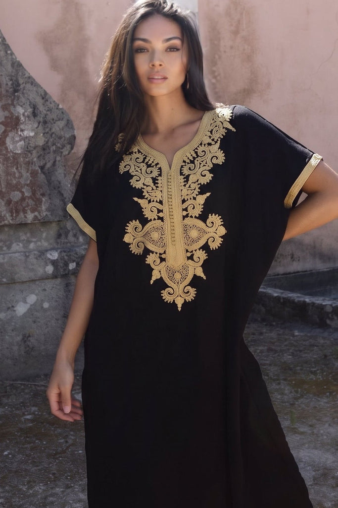 A woman wearing a black caftan dress with ornate gold embroidery around the neckline and sleeve cuffs, standing in front of a rustic pink and gray stone wall.