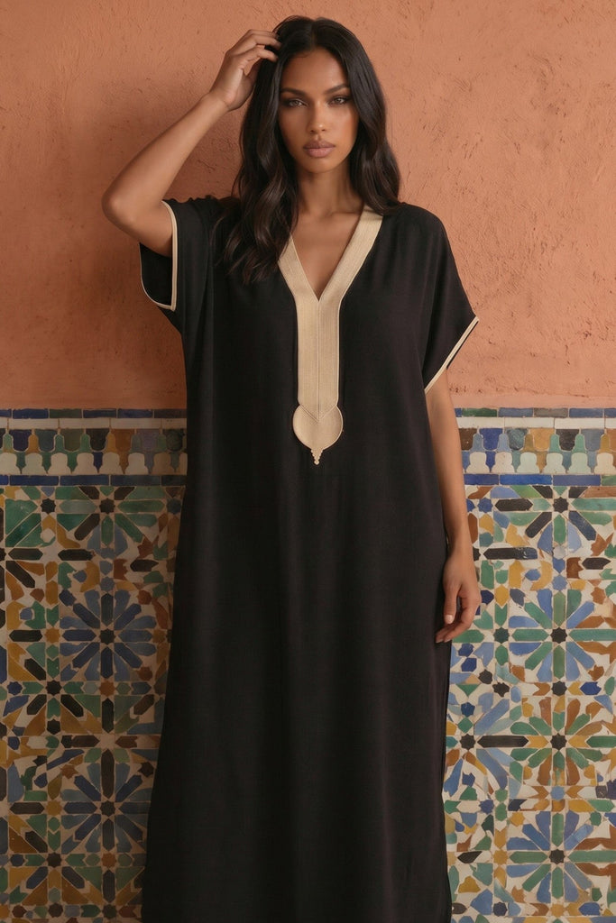 A woman modeling a black kaftan dress with cream-colored trim details, standing in front of a terracotta-colored wall with a traditional patterned tile border.
