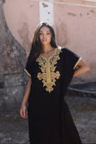 A woman wearing a black caftan dress with ornate gold embroidery around the neckline and sleeve cuffs, standing in front of a rustic pink and gray stone wall.