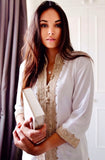 A woman wearing a white long-sleeve tunic with ornate beige embroidery and buttons, holding a white book or box in a softly lit indoor setting with a curtain and white door in the background.