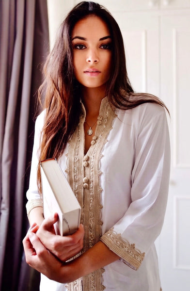 A woman wearing a white long-sleeve tunic with ornate beige embroidery and buttons, holding a white book or box in a softly lit indoor setting with a curtain and white door in the background.