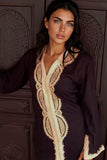 A woman wearing a black long-sleeve tunic with a golden-colored embroidered V-neckline and matching sleeve trim, posed in front of a dark wooden carved door.