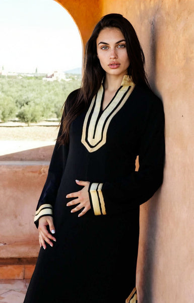 A woman wearing a black long-sleeve abaya with gold and cream geometric trim on the V-neck and cuffs, posing against a warm terracotta archway.