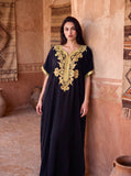 A woman modeling a black kaftan dress with ornate gold embroidery around the neckline and sleeves, standing in a rustic room with terracotta walls and woven textiles.
