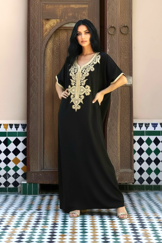 A woman modeling a black kaftan dress with ornate gold embroidery on the neckline, standing in front of a rustic wooden door and patterned tile floor.