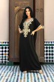 A woman modeling a black kaftan dress with ornate gold embroidery on the neckline, standing in front of a rustic wooden door and patterned tile floor.