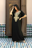 A woman models a black kaftan dress with ornate gold embroidery on the chest and sleeves, standing in front of a traditional wooden door and patterned tile floor.