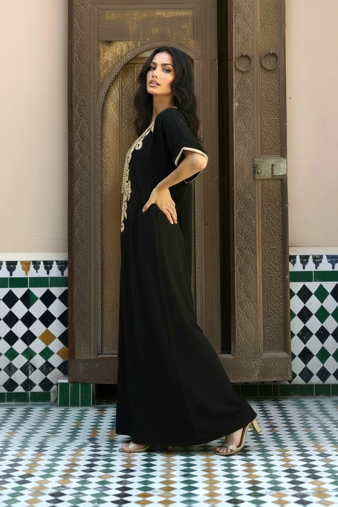 A woman models a black maxi caftan with gold embroidery on a patterned tile floor in front of an ornate wooden door.