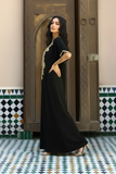 A woman models a black maxi caftan with gold embroidery on a patterned tile floor in front of an ornate wooden door.