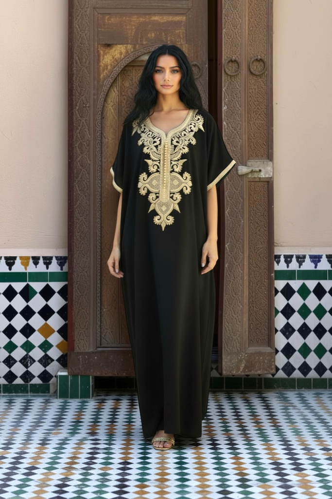 A woman stands in front of a carved wooden door wearing a black caftan with ornate gold embroidery around the neckline and sleeve cuffs. The setting features patterned tile walls and floor.