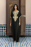A woman stands in front of a carved wooden door wearing a black caftan with ornate gold embroidery around the neckline and sleeve cuffs. The setting features patterned tile walls and floor.