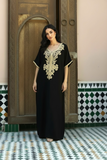 A woman wearing a black caftan with ornate gold embroidery around the neckline, standing in front of a carved wooden door and patterned tile wall.