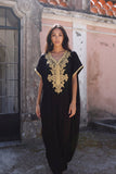 A woman modeling a black caftan dress with ornate gold embroidery on the chest and sleeve cuffs, standing outdoors in front of a rustic stone and plaster building.